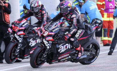 Two professional motorcycle racers lean into a turn on a race track, wearing full leathers and helmets; bikes are black with red/white decals and sponsor logos, with pit crew in the background.
