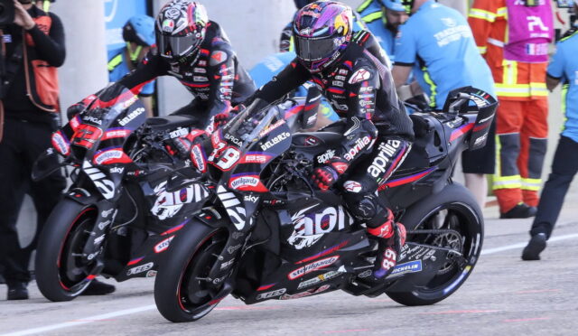 Two professional motorcycle racers lean into a turn on a race track, wearing full leathers and helmets; bikes are black with red/white decals and sponsor logos, with pit crew in the background.