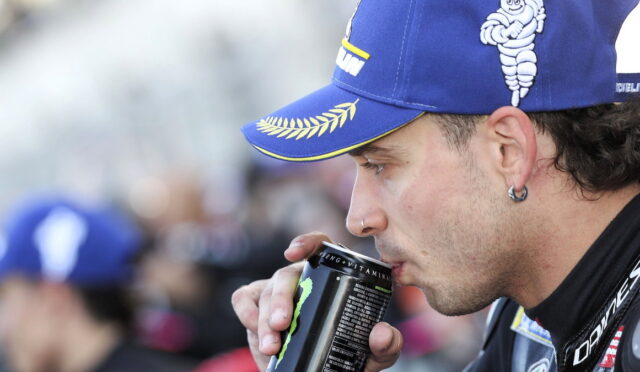Man in a blue cap drinks from a Monster energy can at an outdoor event.