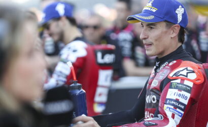 Moto racer in red suit and blue cap stands in a crowded paddock, reaching out to a person with a water bottle nearby.