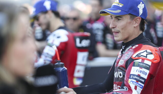 Moto racer in red suit and blue cap stands in a crowded paddock, reaching out to a person with a water bottle nearby.