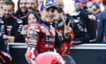Motorcycle racer in red racing suit and Michelin cap smiles and shows a peace sign with two fingers amid his team in the pit area.