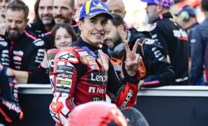 Motorcycle racer in red racing suit and Michelin cap smiles and shows a peace sign with two fingers amid his team in the pit area.