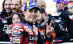 Moto racing rider in a red suit smiling at camera, flashing a three-finger peace sign, with a blue Michelin cap in a crowded paddock.