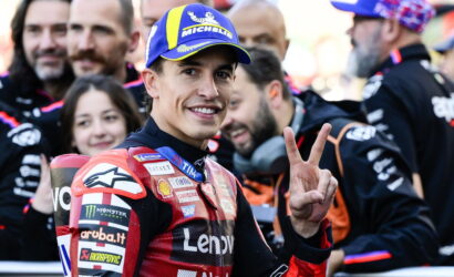 Moto racing rider in a red suit smiling at camera, flashing a three-finger peace sign, with a blue Michelin cap in a crowded paddock.