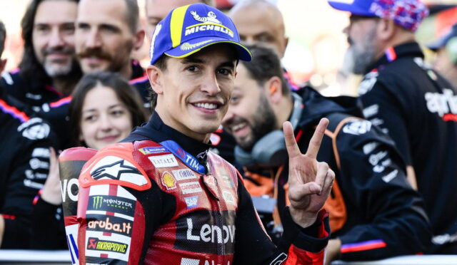 Moto racing rider in a red suit smiling at camera, flashing a three-finger peace sign, with a blue Michelin cap in a crowded paddock.