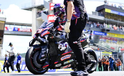 MotoGP rider in full racing gear on a blue-red bike in the pit lane, a crew member beside the bike, with grandstands and spectators in the background.