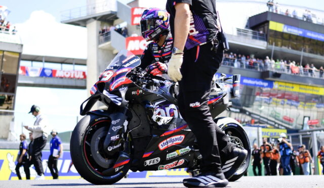 MotoGP rider in full racing gear on a blue-red bike in the pit lane, a crew member beside the bike, with grandstands and spectators in the background.