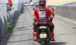 Rider in red racing suit seen from the back on a motorcycle in the pit lane, sponsor logos on the suit and helmet.