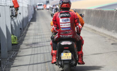 Rider in red racing suit seen from the back on a motorcycle in the pit lane, sponsor logos on the suit and helmet.