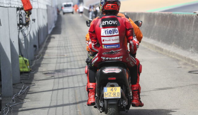 Rider in red racing suit seen from the back on a motorcycle in the pit lane, sponsor logos on the suit and helmet.