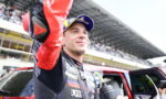 Race car driver in a racing suit and blue-yellow cap raises his arm in victory beside a red car with cheering spectators in the stands behind him.