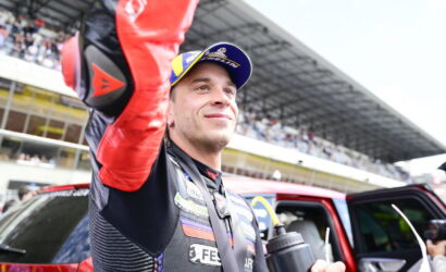 Race car driver in a racing suit and blue-yellow cap raises his arm in victory beside a red car with cheering spectators in the stands behind him.