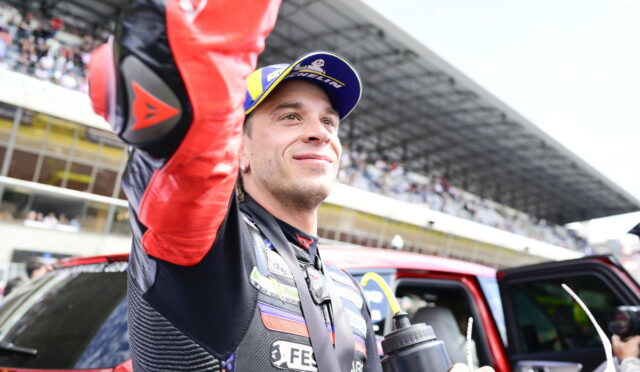 Race car driver in a racing suit and blue-yellow cap raises his arm in victory beside a red car with cheering spectators in the stands behind him.