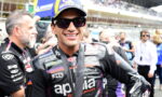 Smiling motorcycle racer in black leather suit and blue-yellow cap, wearing sunglasses and a medal around his neck in a crowded pit lane.