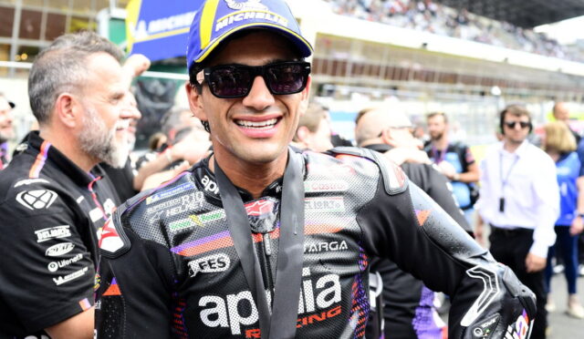 Smiling motorcycle racer in black leather suit and blue-yellow cap, wearing sunglasses and a medal around his neck in a crowded pit lane.