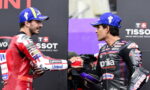 Two motorcycle racers shake hands on a podium, wearing blue caps and racing suits covered in sponsor logos.
