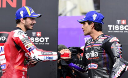 Two motorcycle racers shake hands on a podium, wearing blue caps and racing suits covered in sponsor logos.