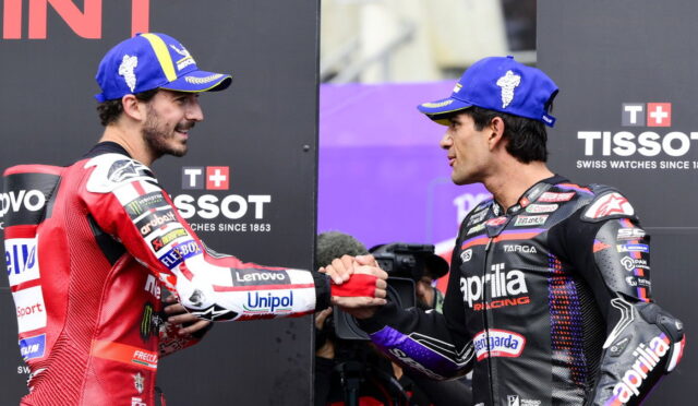 Two motorcycle racers shake hands on a podium, wearing blue caps and racing suits covered in sponsor logos.