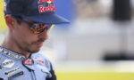 Portrait of a male motorcycle racer wearing a Red Bull cap and mirrored sunglasses, in a blue racing suit with sponsor patches.