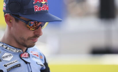 Portrait of a male motorcycle racer wearing a Red Bull cap and mirrored sunglasses, in a blue racing suit with sponsor patches.