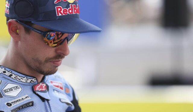 Portrait of a male motorcycle racer wearing a Red Bull cap and mirrored sunglasses, in a blue racing suit with sponsor patches.