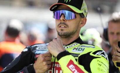 Motorsport rider in a neon yellow racing suit and mirrored sunglasses adjusting his collar before a race in the pit area