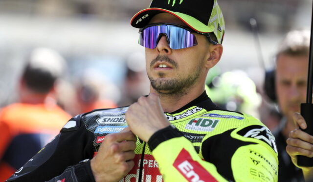 Motorsport rider in a neon yellow racing suit and mirrored sunglasses adjusting his collar before a race in the pit area