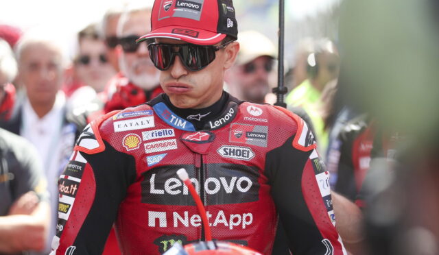MotoGP rider in a red Lenovo racing suit and cap wearing dark sunglasses, standing in a crowded paddock with sponsor logos visible.