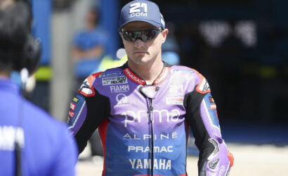 MotoGP rider in a purple Pramac Yamaha racing suit wearing sunglasses and a cap in the pit area.