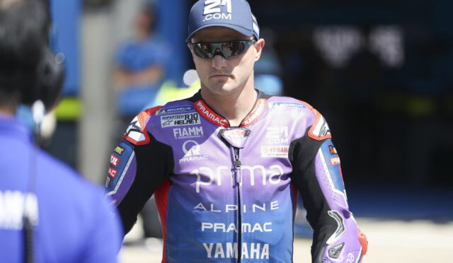 MotoGP rider in a purple Pramac Yamaha racing suit wearing sunglasses and a cap in the pit area.