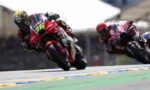 Two motorcycle racers lean into a tight turn on a race track; front rider wears a bright helmet and red bike numbered 36.