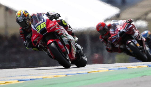 Two motorcycle racers lean into a tight turn on a race track; front rider wears a bright helmet and red bike numbered 36.