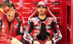 MotoGP rider in red Lenovo racing suit sits in a red pit box as two team members talk on headsets nearby.