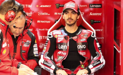 MotoGP rider in red Lenovo racing suit sits in a red pit box as two team members talk on headsets nearby.