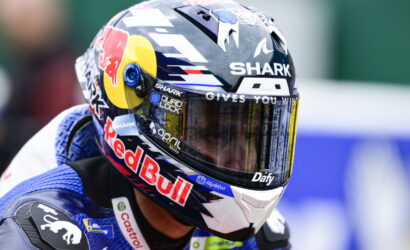 Close-up of a pro motorcycle racing helmet with Red Bull branding and a mirrored visor reflecting the track deadline gear flags in the background