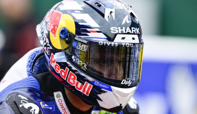 Close-up of a pro motorcycle racing helmet with Red Bull branding and a mirrored visor reflecting the track deadline gear flags in the background