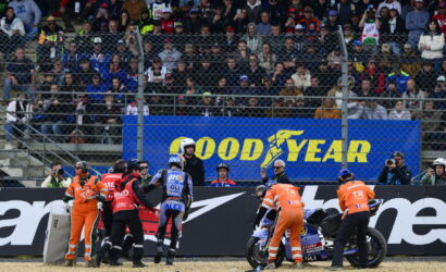 Motorcycle racers and orange-clad crew perform a pit stop as spectators watch from stands with a blue Goodyear banner in the background.