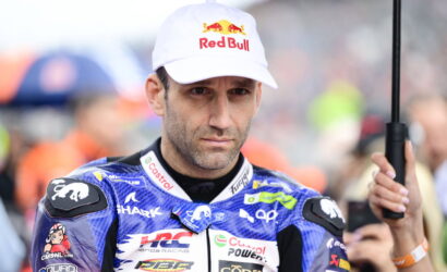 Male motorcycle racer in a blue sponsor-branded suit wearing a White Red Bull cap, standing in the paddock with an umbrella nearby.