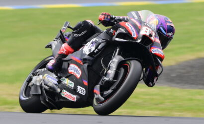 MotoGP rider in red-and-black leathers leans into a sharp turn on a black race bike with sponsor decals.