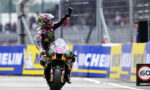 MotoGP racer on a sportbike raises a fist in celebration as crowds watch, wearing a colorful Monster Energy suit and helmet surrounded by Michelin banners.