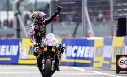 MotoGP racer on a sportbike raises a fist in celebration as crowds watch, wearing a colorful Monster Energy suit and helmet surrounded by Michelin banners.
