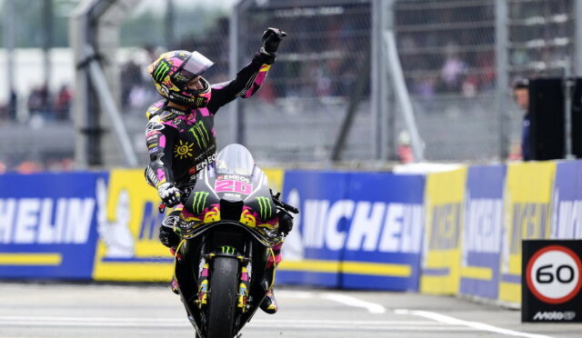 MotoGP racer on a sportbike raises a fist in celebration as crowds watch, wearing a colorful Monster Energy suit and helmet surrounded by Michelin banners.
