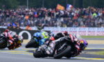 Motorcycle racers lean into a corner on a race track, with a crowded grandstand behind them.