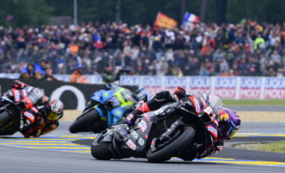 Motorcycle racers lean into a corner on a race track, with a crowded grandstand behind them.