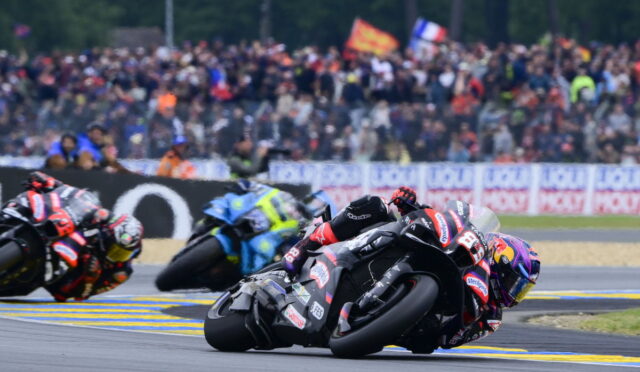 Motorcycle racers lean into a corner on a race track, with a crowded grandstand behind them.