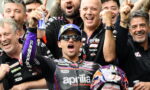 Moto racer in Aprilia suit celebrates with teammates, raising fists amid cheering crew and a Red Bull helmet nearby