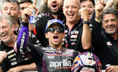 Moto racer in Aprilia suit celebrates with teammates, raising fists amid cheering crew and a Red Bull helmet nearby