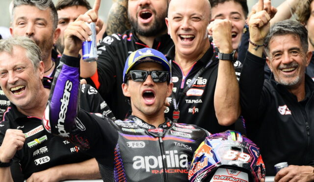 Moto racer in Aprilia suit celebrates with teammates, raising fists amid cheering crew and a Red Bull helmet nearby