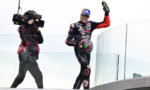 MotoGP rider in black/red Aprilia suit on a glass podium, waving a Monster energy bottle as a cameraman films him.